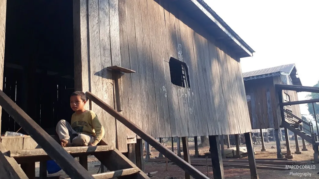 Un bambino di Koh Pek seduto sui gradini d'ingresso della sua casa in legno